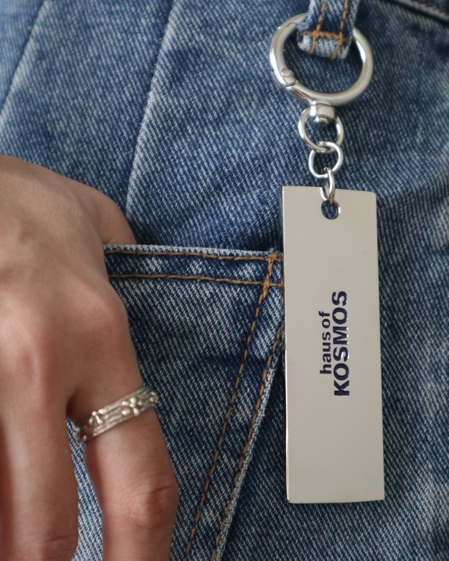 Keychain with engraved 'HAUS OF KOSMOS" logo attached to a denim jeans belt loop.