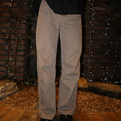 A pair of straight-cut washed denim jeans displayed against an outdoor backdrop with brick wall and foliage in the background.